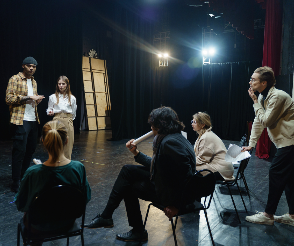 Groep mensen op een theatervloer tijdens een repetitie. Twee acteurs staan tegenover een zittend publiek dat bestaat uit regisseurs en schrijvers met papieren en scripts in hun handen. De ruimte heeft zwarte gordijnen, houten panelen en felle toneelverlichting op de achtergrond.