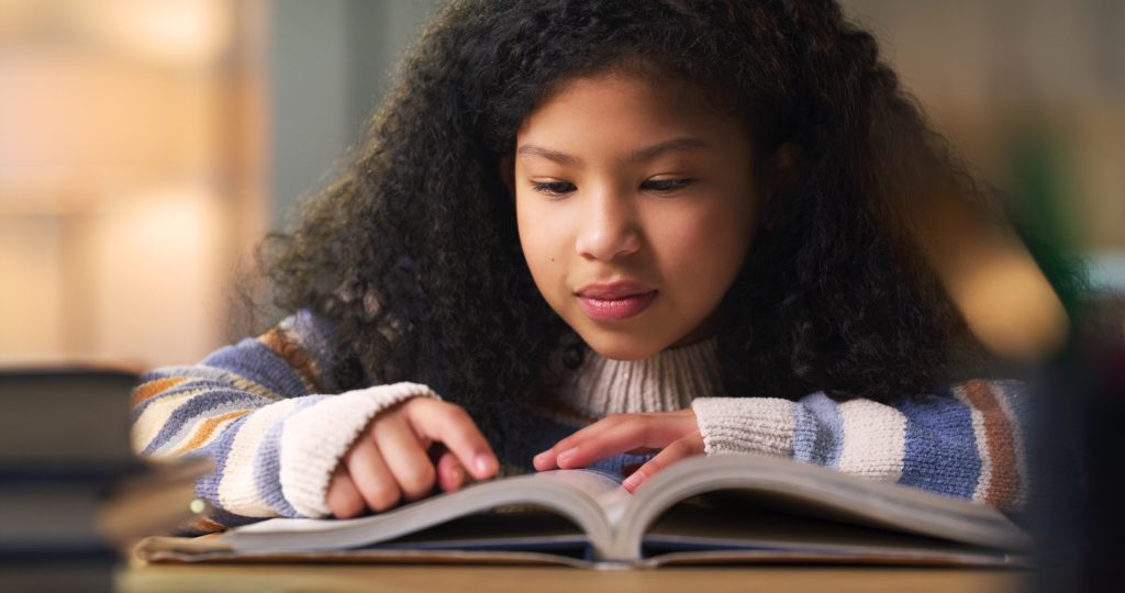 Een jong meisje zit aan tafel en leest een boek. 