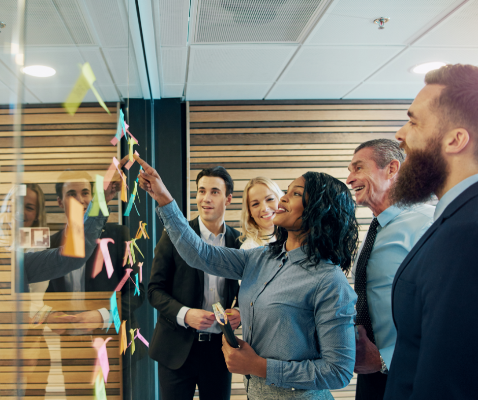 Foto waarop vijf personen in formele kleding te zien zijn. Een van hen wijst naar een met post-its beplakte glazen wand, de rest kijkt ernaar. 