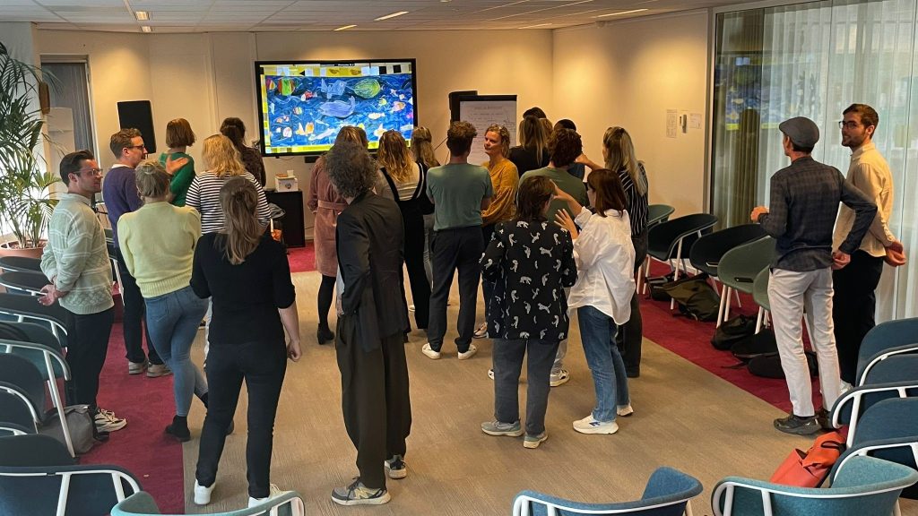 Een groep deelnemers staat in een kring tijdens een training bij LKCA in Utrecht. Op de achtergrond is een groot scherm te zien met kleurrijke kunst. De ruimte heeft stoelen aan de zijkant en een flip-over. 